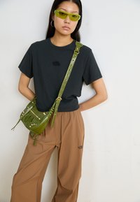 T-shirt noir avec un petit logo, pantalon marron à coupe décontractée et un petit sac en cuir vert avec des accents métalliques, porté en bandoulière avec une sangle.