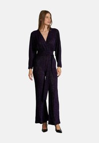 Mono morado con mangas largas, escote en V profundo, tejido texturizado, cintura ceñida y pantalones de pierna ancha, complementado con un cinturón de tela.