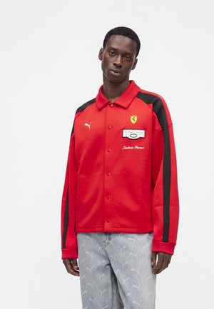 Person trägt eine rote Puma- und Scuderia Ferrari-Jacke mit schwarzen Streifen an den Ärmeln und hellblaue Hosen mit weißen Puma-Logos.