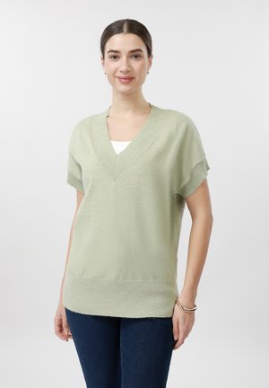 Frau mit zurückgebundenen dunklen Haaren, die einen hellgrünen Kurzarm-V-Ausschnittpullover und dunkelblaue Jeans trägt, steht vor einem schlichten weißen Hintergrund.