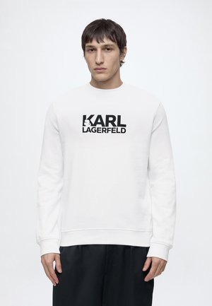Hombre con pelo corto y oscuro que lleva una sudadera blanca con el logo negro de KARL LAGERFELD y pantalones negros, de pie frente a un fondo blanco liso.