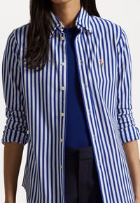 Blå och vit randig skjorta med knappar och krage, med en enda logotypaccent, rullade ärmar och en lätt tygtextur.