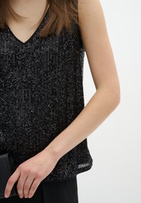 Top nero in paillettes con scollatura a V, caratterizzato da una vestibilità loose e una superficie lucida e texturizzata, abbinato a una clutch nera in mano.