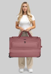 Joven con cabello largo y rubio sosteniendo una gran bolsa de viaje con ruedas en color malva, vistiendo una camisa blanca, pantalones beige y zapatillas blancas sobre un fondo gris.
