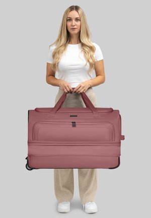 Giovane donna con lunghi capelli biondi che tiene una grande borsa a rotelle mauve, indossando una camicia bianca, pantaloni beige e sneakers bianche su sfondo grigio.