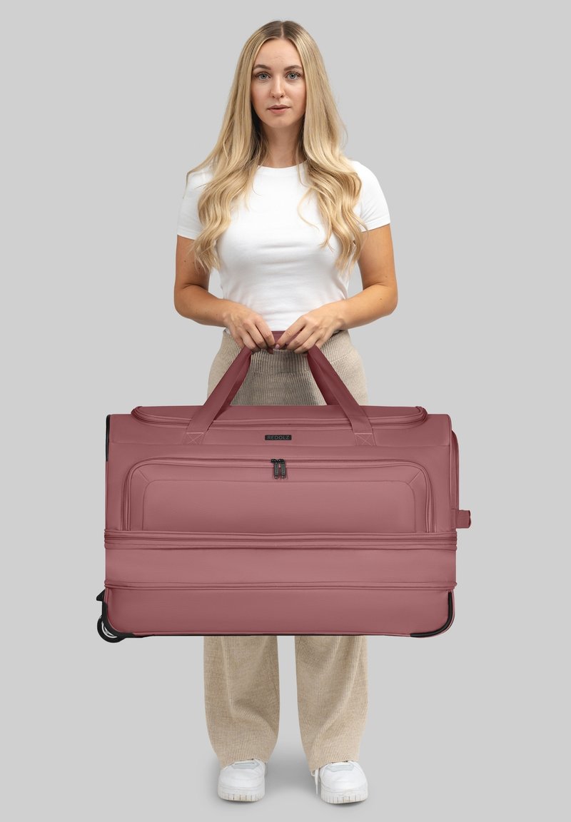 Joven con cabello largo y rubio sosteniendo una gran bolsa de viaje con ruedas en color malva, vistiendo una camisa blanca, pantalones beige y zapatillas blancas sobre un fondo gris.