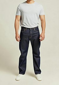 Homme portant un t-shirt blanc uni, un jean droit bleu marine et des baskets blanches, debout devant un fond neutre.