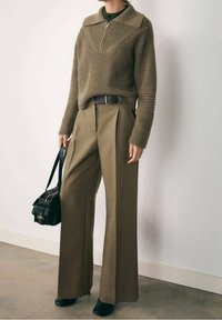 Olivově zelený žebrovaný pletený svetr s polovičním zipem u límce; široké béžové kalhoty; černá kabelka přes rameno; robustní černé boty.
