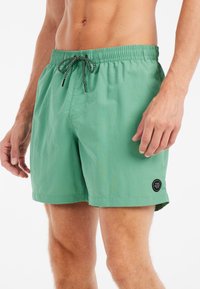 Shorts de bain vert clair avec une ceinture élastique, un cordon noir, une poche latérale et un patch rond noir sur le bas de la jambe gauche.