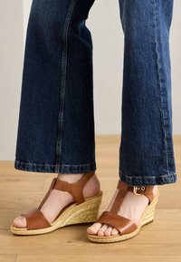 Brown leather wedge sandals with a woven jute outsole, open toe design, and ankle strap with buckle detail, paired with blue denim trousers.
