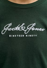Tume roheline dressipluus, mille esiküljel on valge tikandiga tekst "Jack & Jones" ja "Nineteen Ninety" lõbusas fondis.