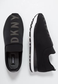 Baskets DKNY noires à enfiler en tissu lisse, arborant un logo en gris foncé et une semelle blanche avec une adhérence texturée.