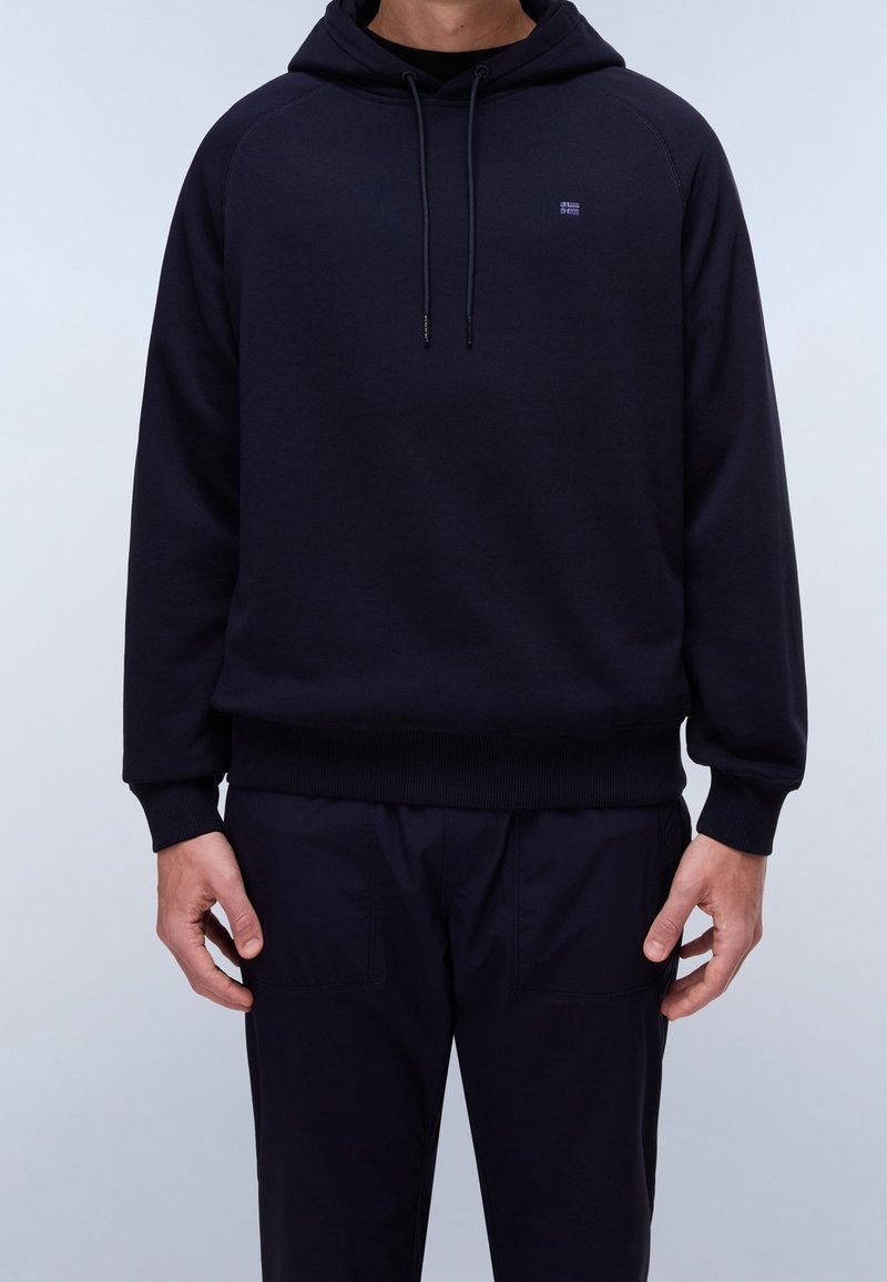 Personne portant un sweat à capuche bleu marine uni avec cordons et un pantalon bleu marine assorti, debout, les mains détendues le long du corps.