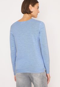 Helleblaues langärmliges Sweatshirt mit einem strukturierten Muster, weitem Ausschnitt und leicht abgerundetem Rückensaum. Getragen mit grauen Hosen.