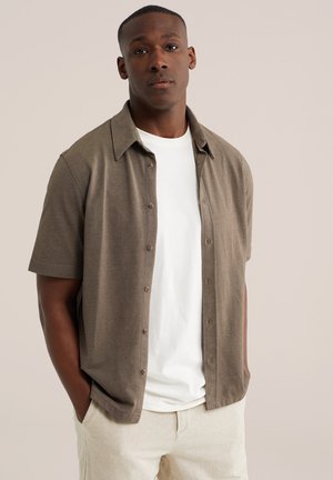 Homme debout les mains dans les poches, portant une chemise marron à manches courtes boutonnée sur un t-shirt blanc et un pantalon beige clair sur fond neutre.