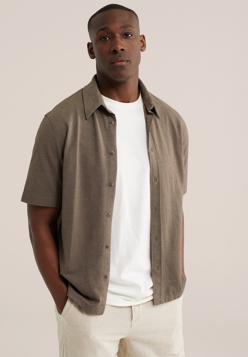 Mann steht mit den Händen in den Taschen und trägt ein braunes kurzärmeliges Hemd mit Knöpfen über einem weißen T-Shirt sowie hellbeige Hose vor neutralem Hintergrund.