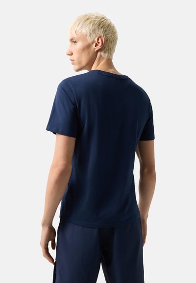 Jeune personne aux cheveux blonds courts portant un t-shirt bleu marine uni et un pantalon assorti, vue de dos avec la tête tournée vers la gauche.