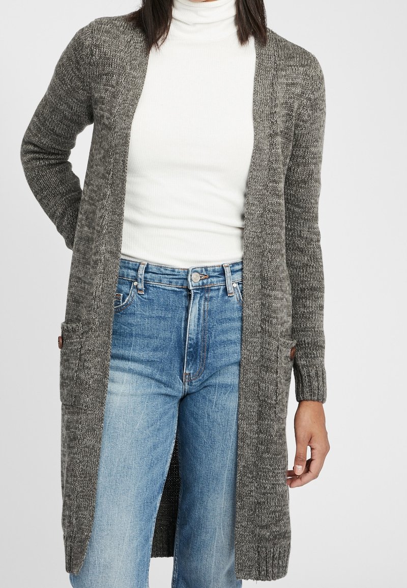 Femme portant un long cardigan gris en tricot par-dessus un col roulé blanc et un jean taille haute bleu clair, debout devant un fond uni.