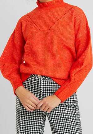 Vrouw in een feloranje, gestructureerde gebreide trui en zwart-wit houndstooth patroonbroek, met de handen op de taille.