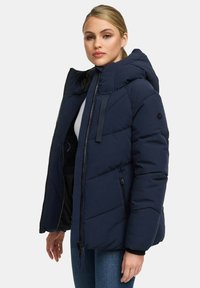 Vrouw in een marineblauwe gewatteerde hooded jacket met ritszakken, staande tegen een effen lichte achtergrond, met een lichte draai naar links.
