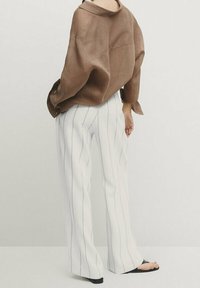 Chemise oversize en lin marron à manches larges associée à un pantalon évasé blanc à rayures. Des sandales noires complètent la tenue.