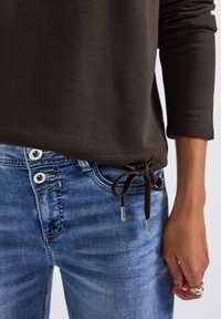 Sweatshirt marron foncé avec un cordon de serrage à l'ourlet et des embouts en métal, associé à un jean déchiré bleu clair avec des boutons blancs.