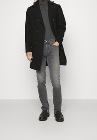 Abrigo negro de lana con doble botonadura sobre un jersey de cuello alto gris, combinado con vaqueros grises y zapatos negros sin cordones. Texturas lisas y diseño ajustado.