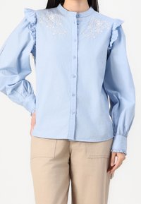 Camicia azzurra chiara con maniche a frangia e dettagli floreali ricamati sul petto. Realizzata in tessuto morbido e leggero.