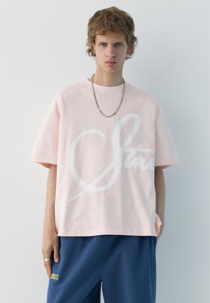 Fiatal férfi göndör hajjal, világos rózsaszín, túlméretezett pólóban, fehér felirattal és tengerészkék nadrágban, sima fehér háttér előtt.