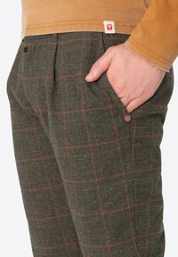 Grüne kariertete Hose mit roten Linien, strukturierter Stoff, seitliche Tasche und Logo-Tag am Bund. Flacher Schnitt mit Falten.