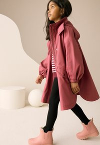 Málnaszínű esőkabát kapucnival, rugalmas mandzsettákkal és patentgombokkal, csíkos felső és fekete leggings fölött viselve, rózsaszín gumicsizmával párosítva.