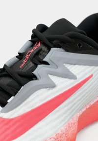 Shoe atlético em preto e branco com um acento rosa, apresentando tecido texturizado, um cabedal em malha respirável e sobreposições sintéticas cinzas.