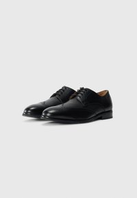 Chaussures de ville pour hommes en cuir noir avec des détails brogues et des lacets, présentées côte à côte sur un fond blanc.