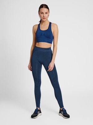 Kvinde står med hånd på låret iført matchende navyblå sports-bh og leggings med sorte sneakers mod en ensfarvet hvid baggrund.
