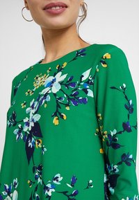 Green floral-patterned dress with a round neckline, featuring blue, white, and yellow flowers on a smooth fabric in a fitted design.