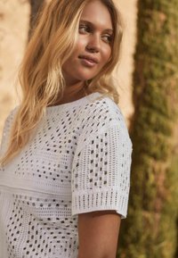 Top bianco lavorato a maglia con un motivo a trama aperta, maniche corte e scollatura arrotondata, ambientato in un contesto naturale all'aperto.