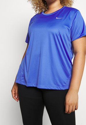 Persona de cabello rizado que lleva una camiseta azul holgada de Nike y pantalones negros, de pie frente a un fondo blanco.