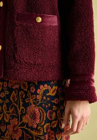 Burgundi, tekstuuriga fliiskinnas, millel on tasku ja kuldne nööpide detail, koos lilli musteriga tume seelikuga, millel on erksad värvid.