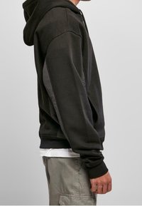 Vista lateral de una persona que lleva una sudadera negra sobre una camisa blanca y pantalones cargo grises claros, contra un fondo claro y simple.