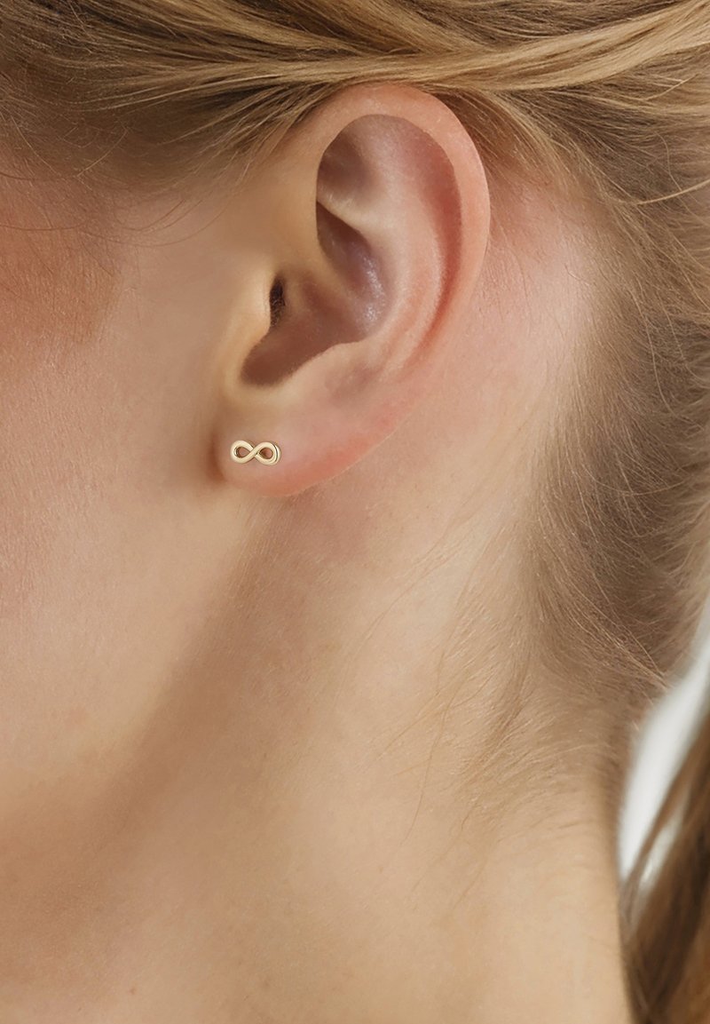 Gros plan sur l'oreille d'une personne portant une petite boucle d'oreille clou en forme de symbole infini en or.