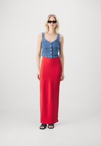 Haut en denim à manches courtes avec des boutons, associé à une longue jupe rouge à pois. Le mannequin porte des chaussures noires à bout ouvert et des lunettes de soleil sombres.