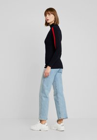 Marineblauwe turtleneck trui met rode strepen op de mouwen, gecombineerd met lichtblauwe jeans en witte sneakers, met een slim fit ontwerp.