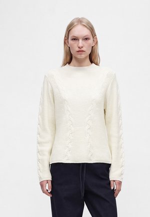 Femme portant un pull en maille torsadée crème à manches longues et un pantalon foncé, debout devant un fond uni clair.