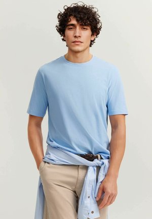 Young man with curly hair wearing a light blue t-shirt and beige pants, with a blue striped shirt tied around his waist.