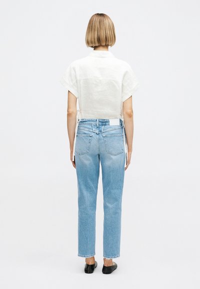 Personne aux cheveux blonds courts portant une chemise blanche à manches courtes, un jean droit bleu clair et des chaussures noires sans lacets, debout de dos.