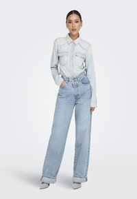 Camisa de denim azul claro con dos bolsillos en el pecho, combinada con jeans de cintura alta y pierna ancha en azul claro y tacones de punta en azul claro.