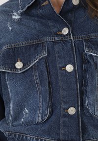 Denim jacket met een donkerblauwe kleur, versleten details, twee borstzakken en zilveren drukknoopjes aan de voorkant.
