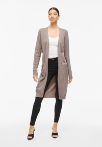 Kvinne iført en lang lilla cardigan over en hvit topp, svarte skinny jeans og svarte åpne hæler, står foran en hvit bakgrunn.