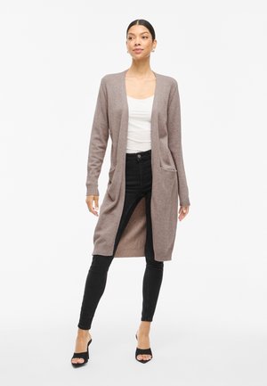 Frau in einem langen mauvefarbenen Cardigan über einem weißen Oberteil, schwarzen engen Jeans und schwarzen Open-Toe-Highheels, die vor einem weißen Hintergrund steht.