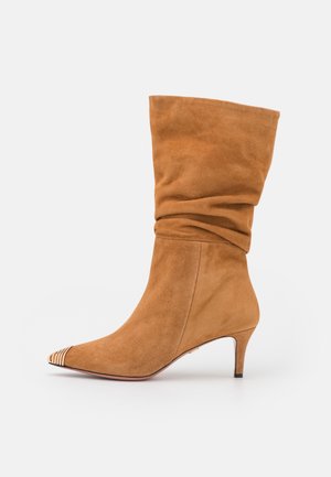 Zamszowe botki za kostkę w jasnobrązowym kolorze, z luźnym cholewką, spiczastym noskiem i dekoracyjnym paskiem wzdłuż noska; obcas o wysokości 3 cali.